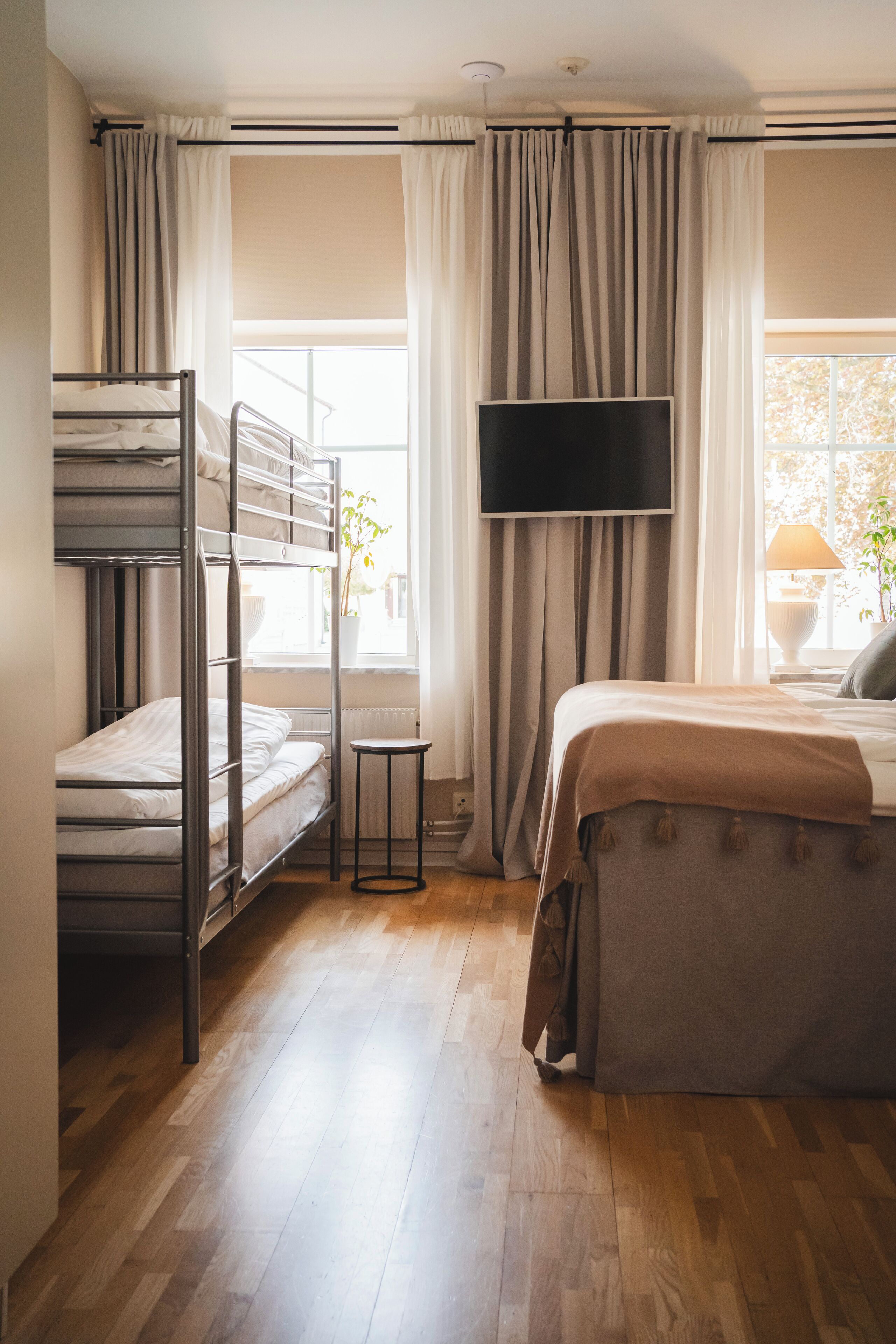 A hotel room with a bunk bed, a single bed, a flat-screen TV, a small table, and a window with curtains.