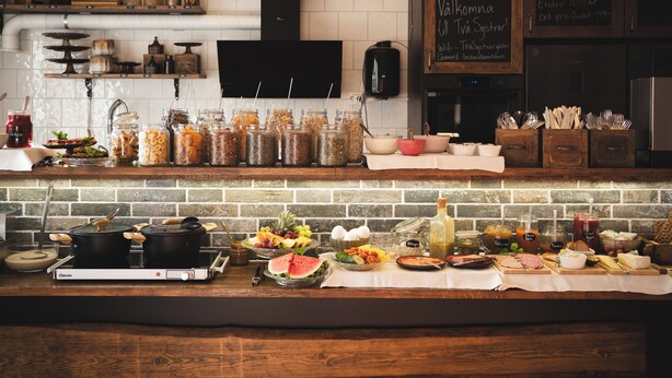Una encimera de cocina con diversos alimentos y utensilios de cocina. Hay frascos con alimentos en conserva, un bol de fruta y una botella de salsa.