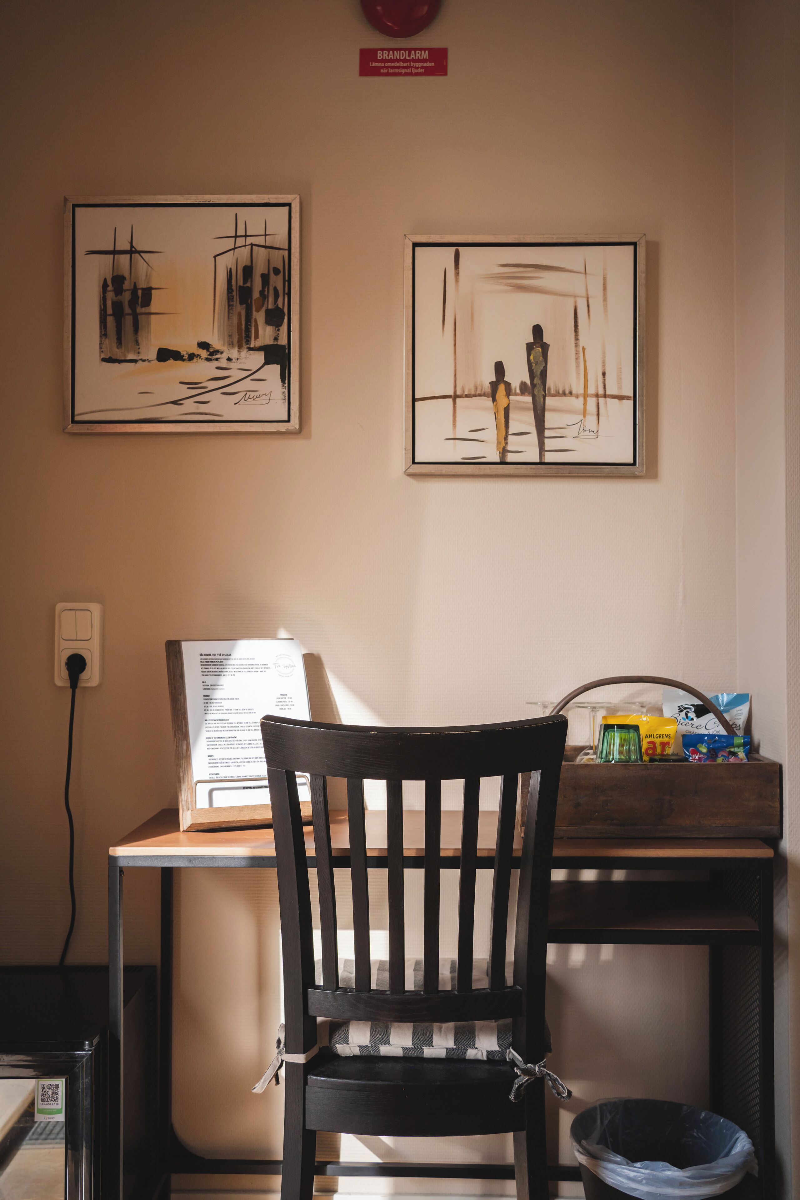 A room with a wooden chair, a table with a laptop, and two framed artworks on the wall.