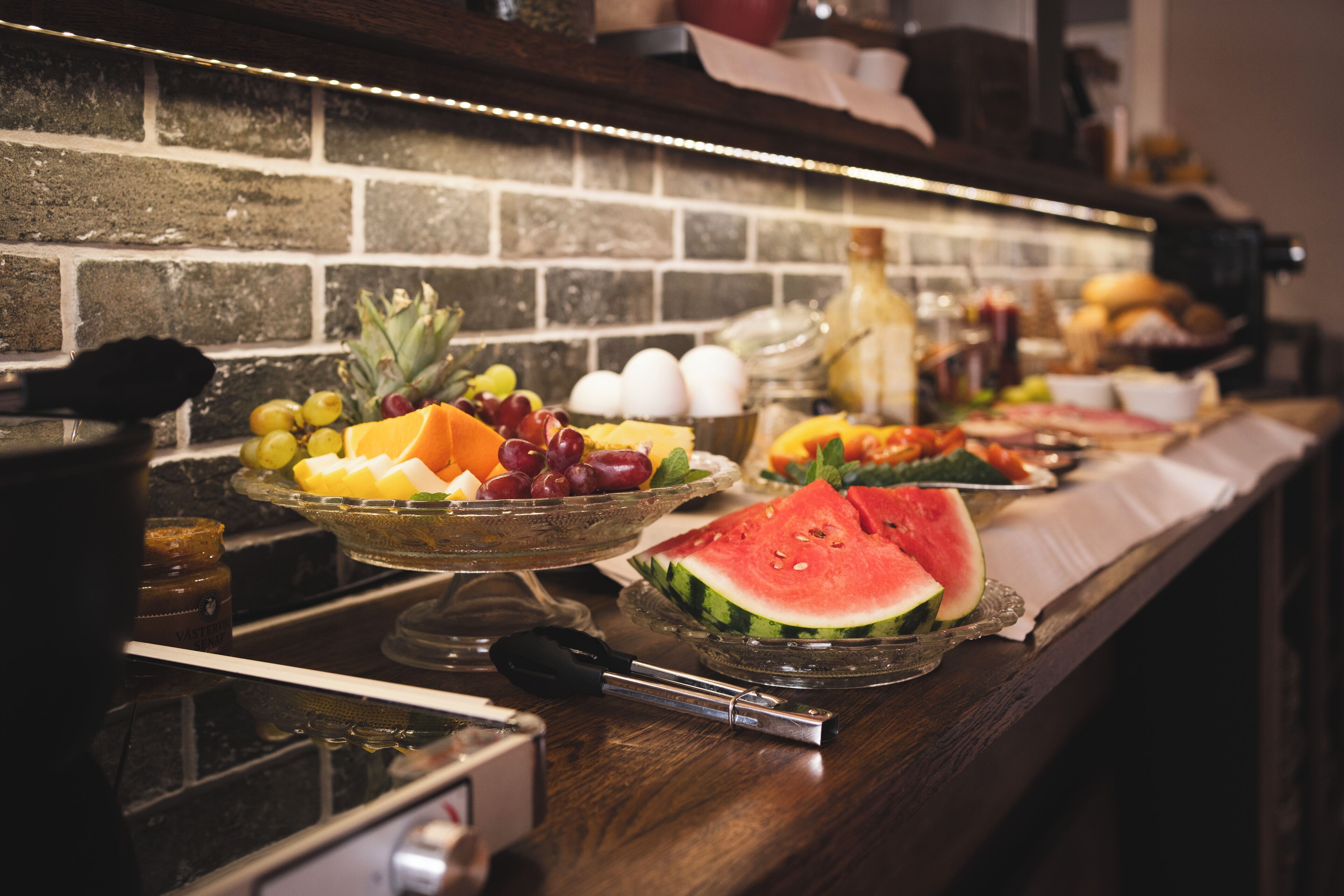 Un buffet proposant une variété de fruits et de légumes, notamment des pastèques et des raisins, sur un comptoir en bois avec un mur de briques en arrière-plan.
