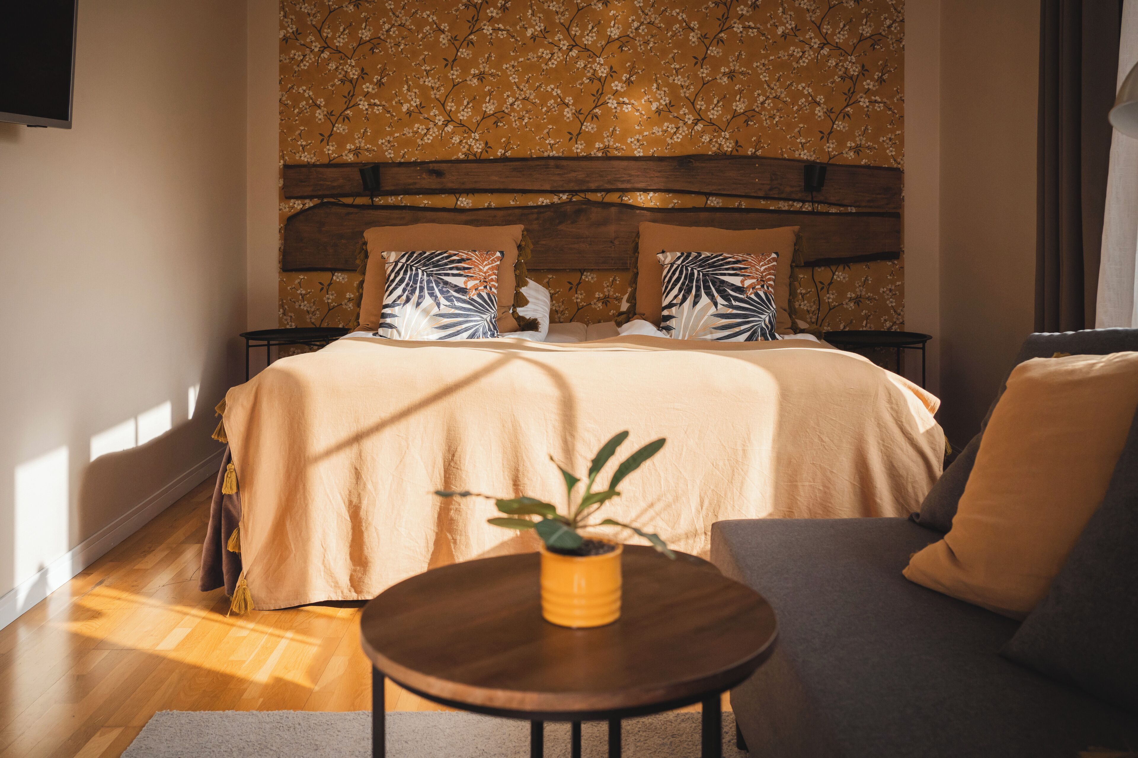 A bedroom with a wooden headboard, floral wallpaper, a bed with beige bedding, and a small round table with a plant.