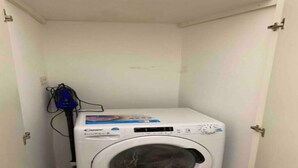 A compact, white washing machine in a small, enclosed laundry room with a door on one side and a power outlet on the wall. - Il loft del vico - Salento (galatone)
