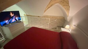A bedroom with a red bedspread, a television displaying a fire, and a stone wall. - Il loft del vico - Salento (galatone)
