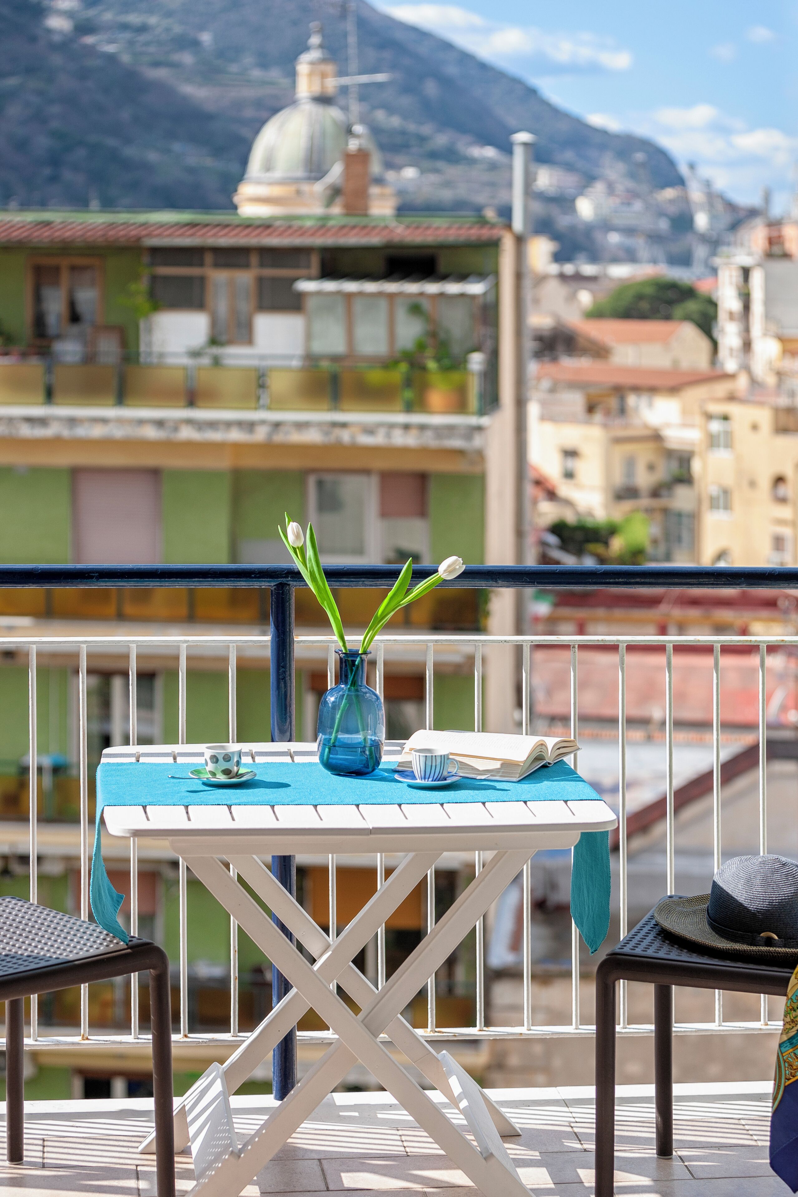 A balcony with a table, chairs, a vase with flowers, and a view of buildings and a mountain in the background.