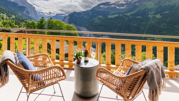 A balcony with wicker chairs, a table with a bottle and glasses, and a view of mountains. - Chalet Alpenheim 1 (Lauterbrunnen)
