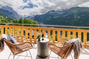 A balcony with wicker chairs, a table with a bottle and glasses, and a view of mountains. - Chalet Alpenheim 1 (Lauterbrunnen)