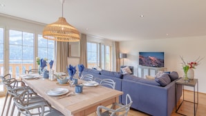 A modern living room with a wooden dining table, grey chairs, a blue sofa, and a large window offering a mountain view. - Chalet Alpenheim 1 (Lauterbrunnen)
