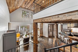 A spacious loft-style interior with a high ceiling, exposed wooden beams, a kitchen with bar stools, and a dining area with a large table and chairs. - Row House | Lakefront Luxury, Private Dock, Gas Grill & Fire Pit! (Nebo)