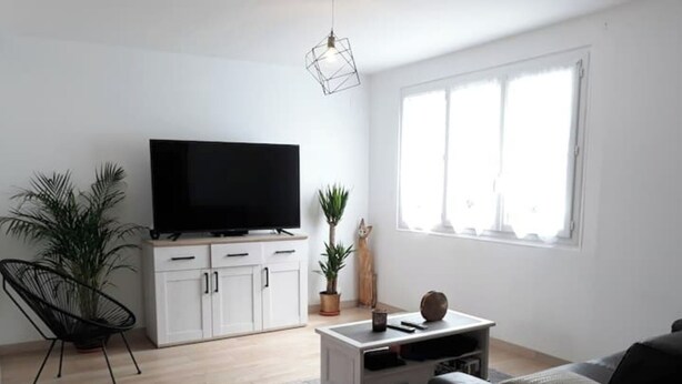 A modern living room with a flat-screen TV on a white cabinet, a black chair, a coffee table, and a large window providing natural light. - Air-conditioned house 3 min from Carcassonne, swimming pool (Pennautier)