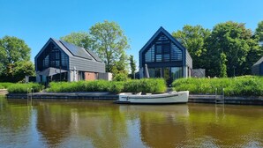 Uma casa-barco moderna com uma pequena varanda e área coberta, atracada em um canal, com vegetação exuberante e céu azul claro ao fundo.