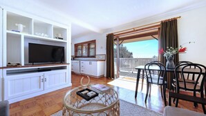 A living room with a TV, white cabinets, a glass coffee table, and a balcony with a dining area. - What A View - Lamont St Unit 4 120 (Bermagui)