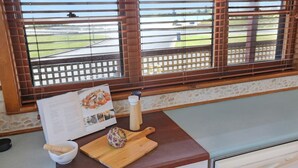 A room with a view of a road and greenery through wooden blinds, a wooden table with a menu, a bowl, a wooden board, and a pepper grinder. - What A View - Lamont St Unit 4 120 (Bermagui)