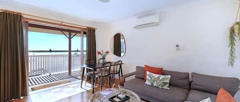 A living room with a sofa, coffee table, and dining area with chairs. There is a view of the sea through large windows.