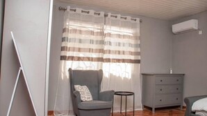 Une pièce avec un fauteuil gris, une petite table ronde, une commode grise et une fenêtre avec des rideaux à rayures.