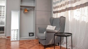 Une pièce comprenant un fauteuil gris, une petite table d’appoint noire, un coussin blanc, une étagère grise et une fenêtre ornée de rideaux légers.