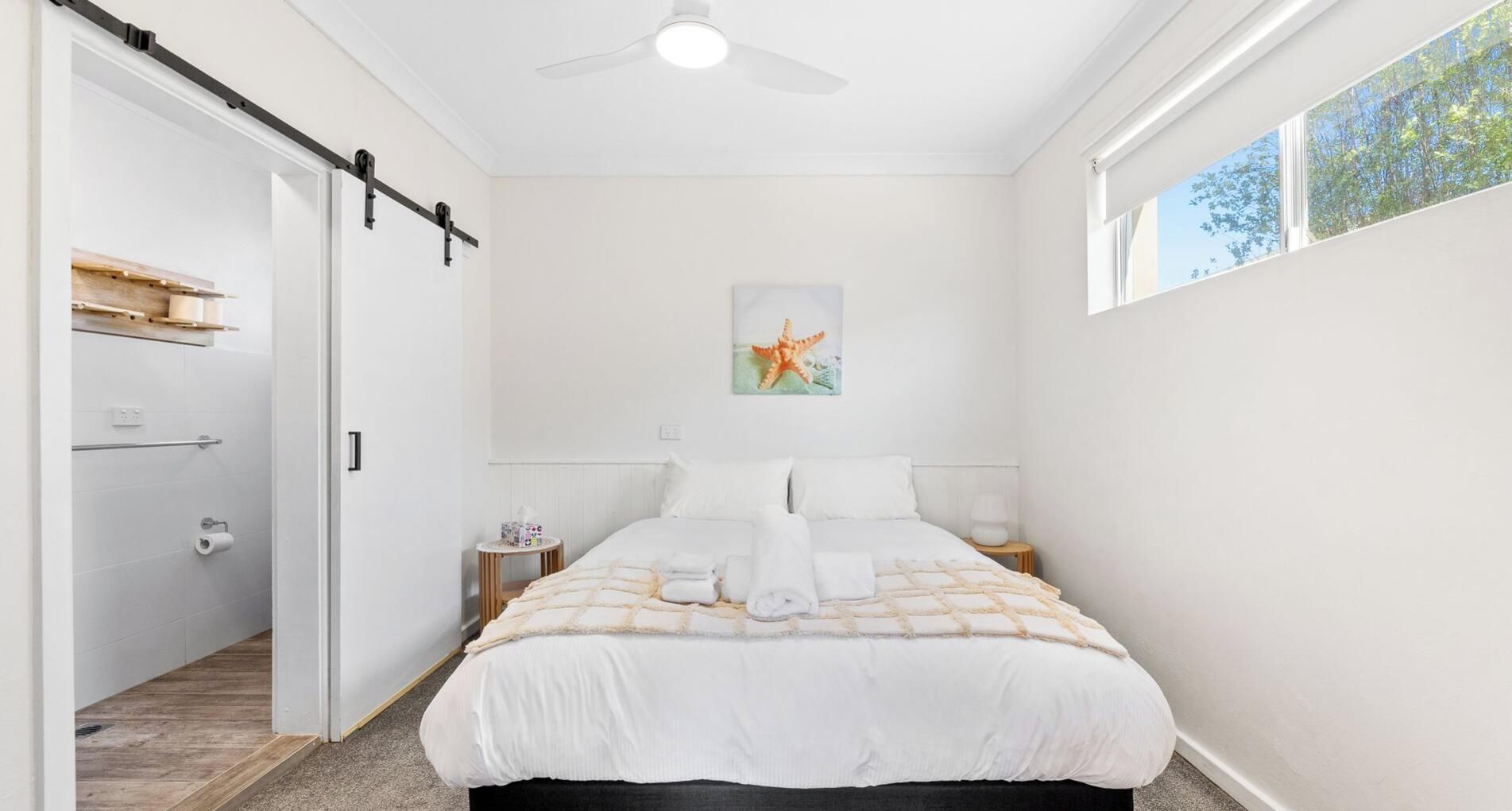 Une chambre à coucher avec un lit, une table de chevet, un tableau, un ventilateur de plafond et une fenêtre.