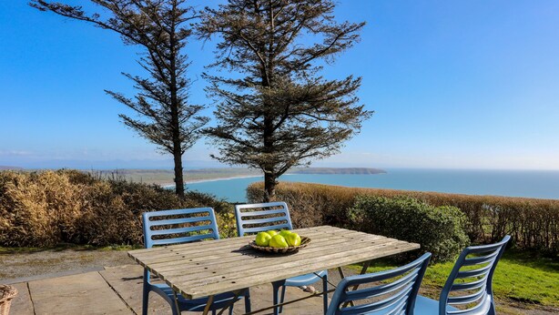 A wooden table with a basket of green apples, surrounded by blue chairs, set on a patio with a view of the sea and hills. - Bryn Meillion (Pwllheli)