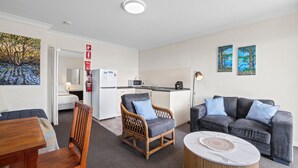 Une chambre d’hôtel compacte comprenant un lit, une table à manger, une chaise en osier, un canapé, un petit coin cuisine et des œuvres d’art accrochées aux murs.