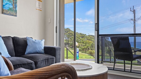 A modern living room with a black sofa, a round coffee table, and a view of a park and ocean through large glass doors.