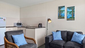 Quarto de hotel compacto com cozinha compacta, área de refeições e sofá com almofadas azuis. Há duas pinturas na parede.