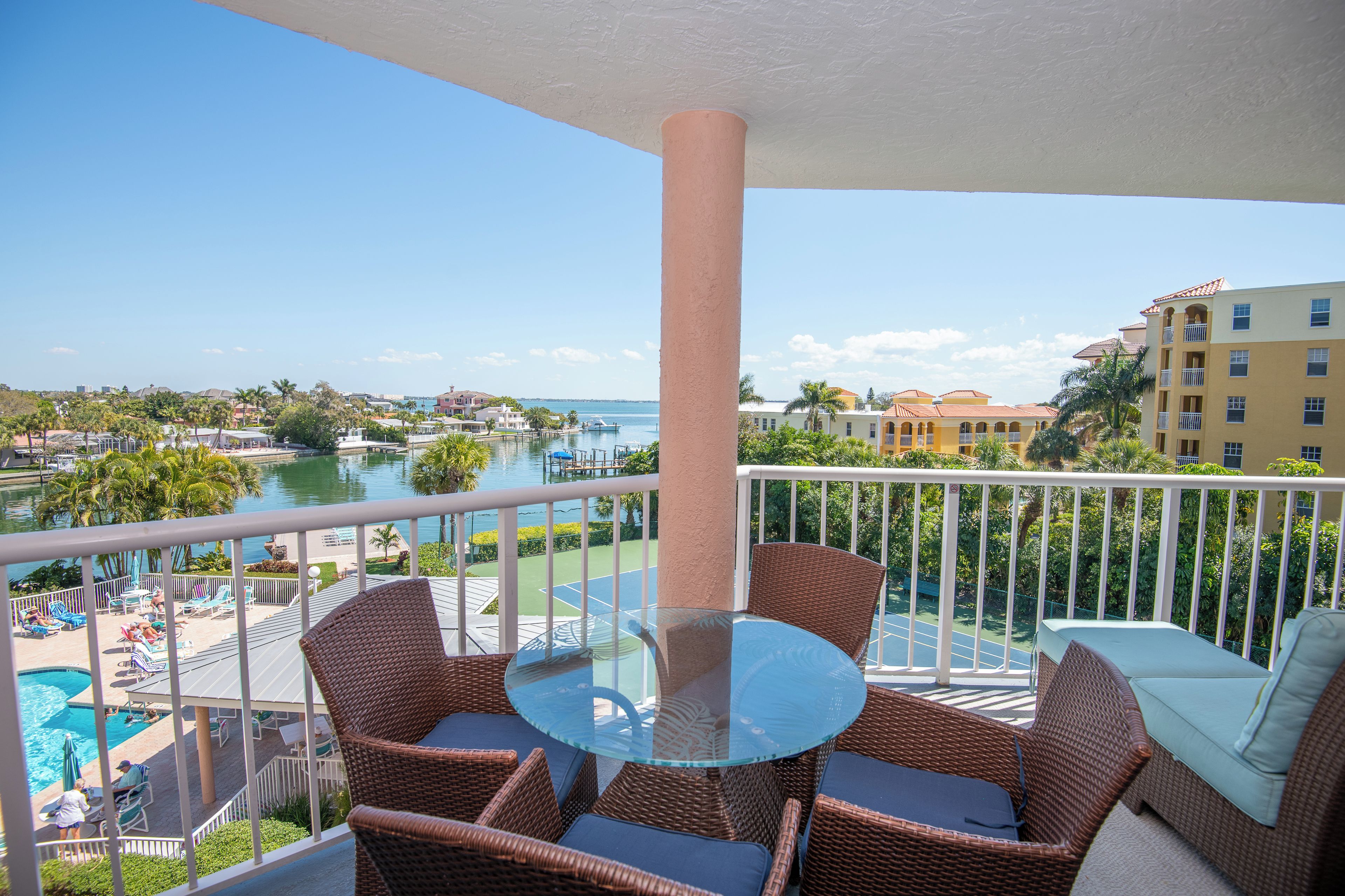 A balcony with wicker furniture, a glass table, and a view of a pool and buildings.