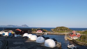 A coastal harbor with several red buildings, a dock with boats, and a clear view of the sea and mountains in the background. - Enjoy Lofoten Glamping (Vestvågøy)