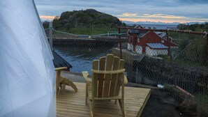 A wooden deck with a chair, a tent, and a view of a body of water and buildings in the background. - Enjoy Lofoten Glamping (Vestvågøy)