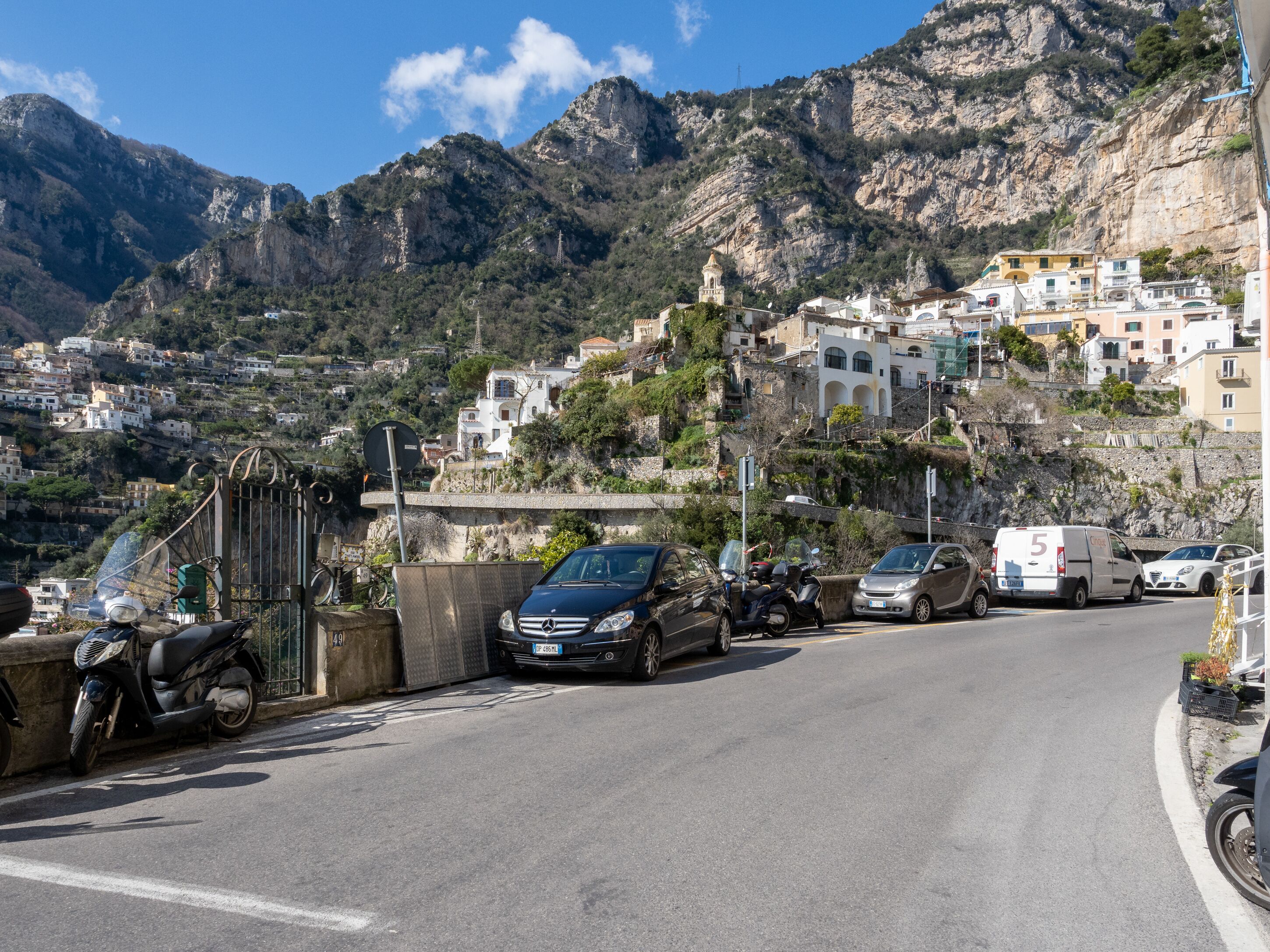 A coastal road with parked cars and a scooter, leading to a hillside with buildings.