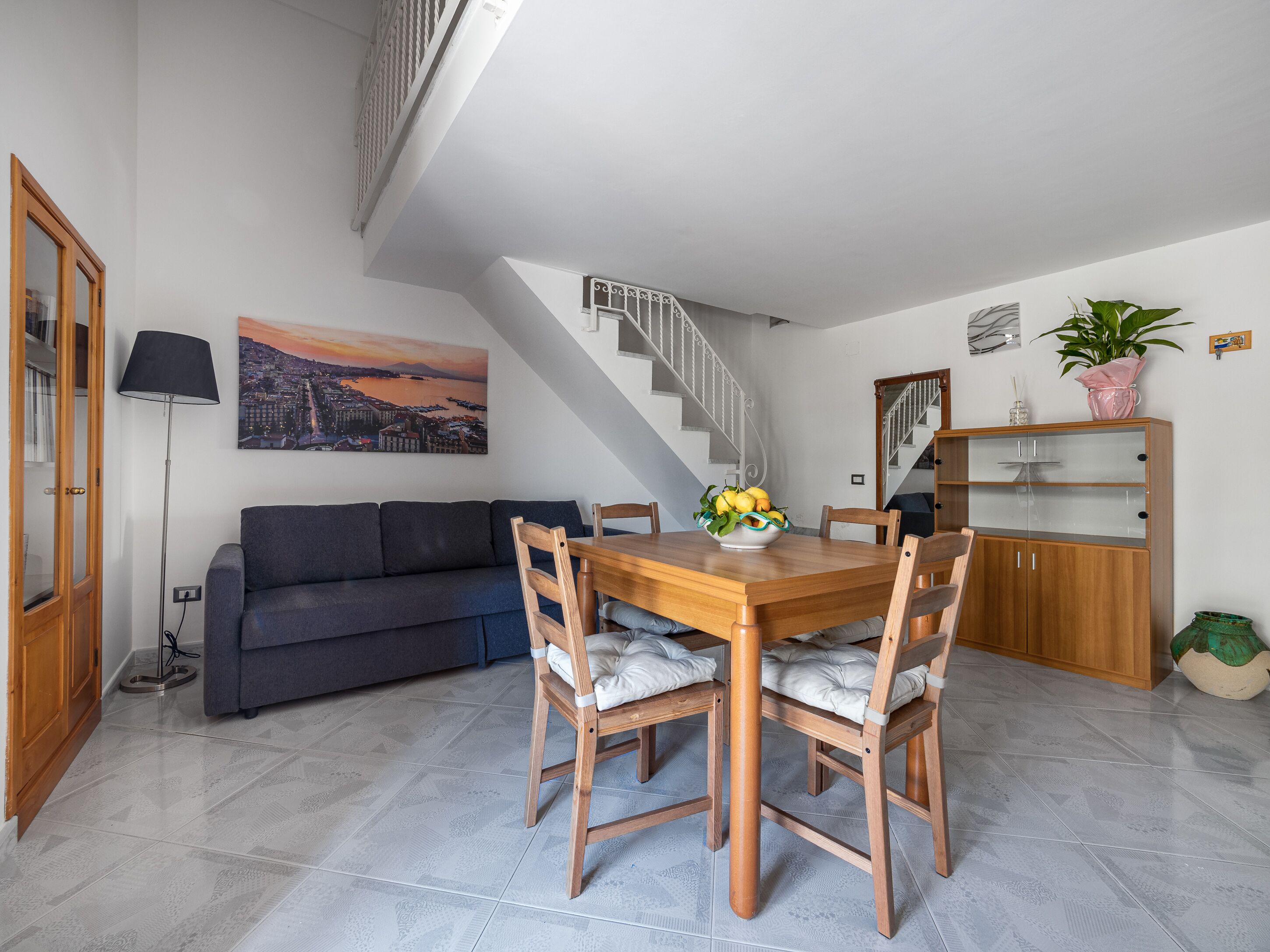 Un coin repas avec une table et des chaises en bois, un canapé, un escalier et un tableau représentant un paysage urbain.