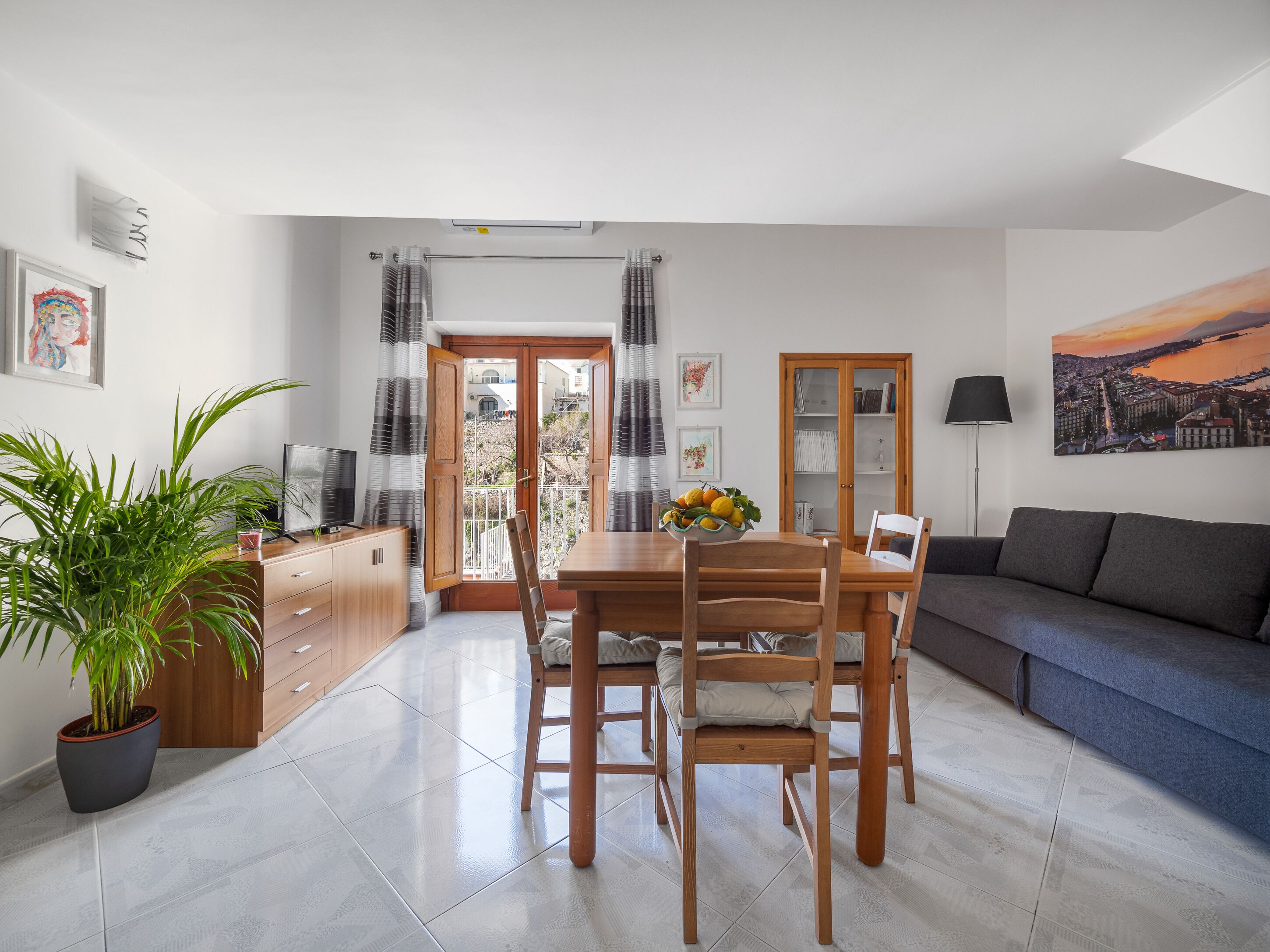 Un salon moderne avec un coin repas, comprenant une table en bois, des chaises, un canapé gris, une plante en pot et un tableau représentant un paysage urbain.