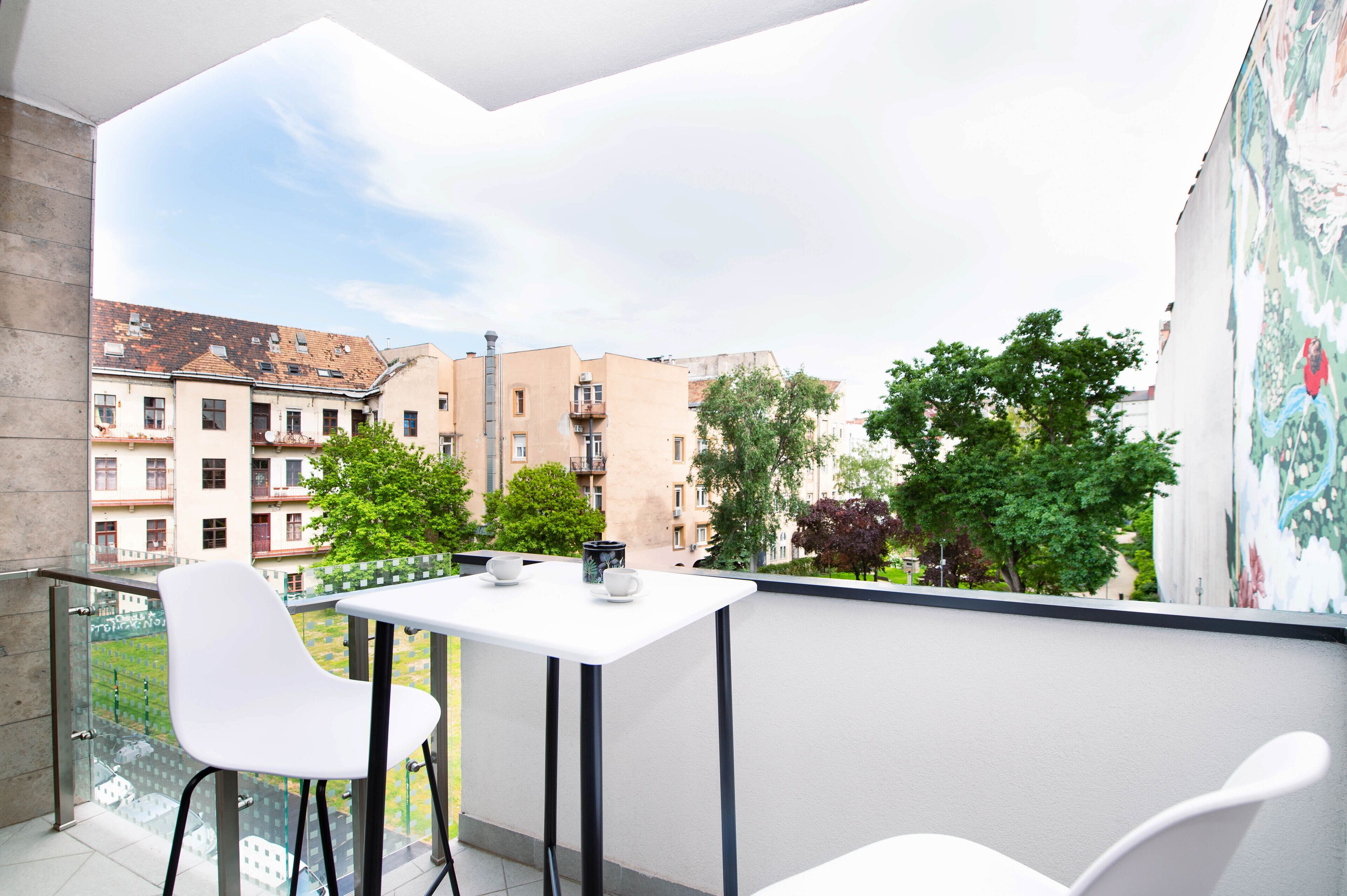 A balcony with a white table and chairs, overlooking a courtyard with buildings and trees.