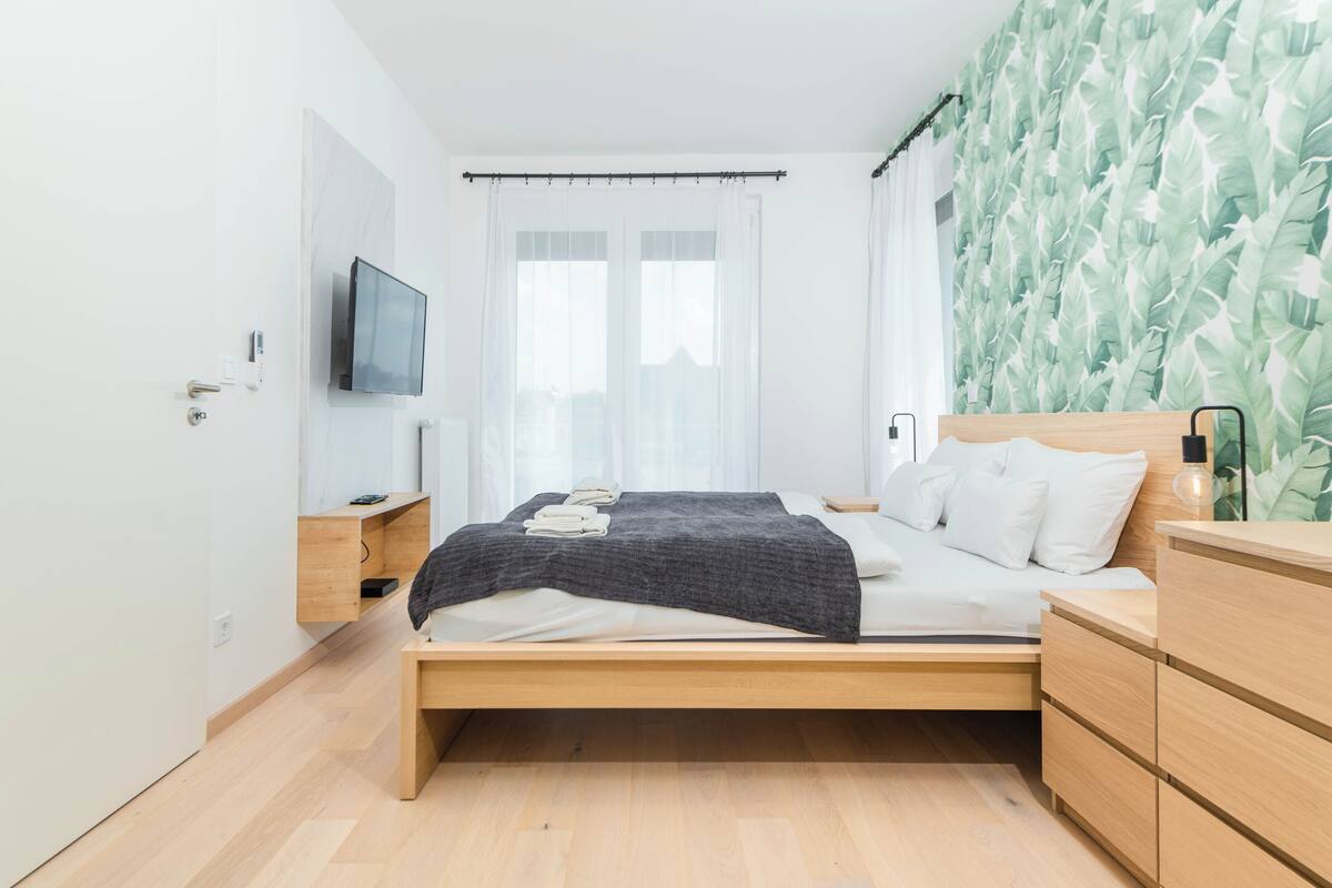 A modern bedroom with a wooden bed, a bedside table, a nightstand, a TV mounted on the wall, and a window with sheer curtains.