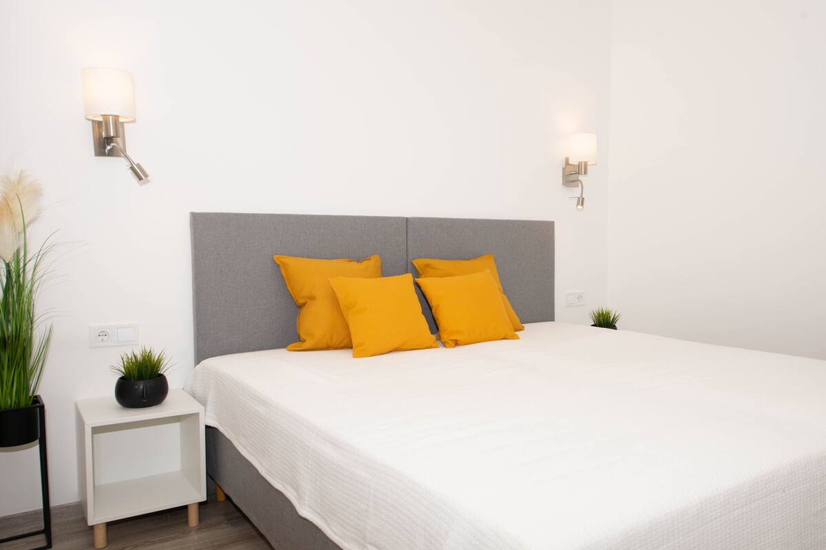 A neatly made bed with a white bedspread and two bright yellow pillows, a small bedside table with a plant, and two wall-mounted light fixtures.