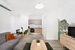 Una sala moderna con un sofá gris, una mesa de centro de madera y un televisor sobre un mueble de madera. Se ve un comedor con una mesa blanca y sillas verdes al fondo.