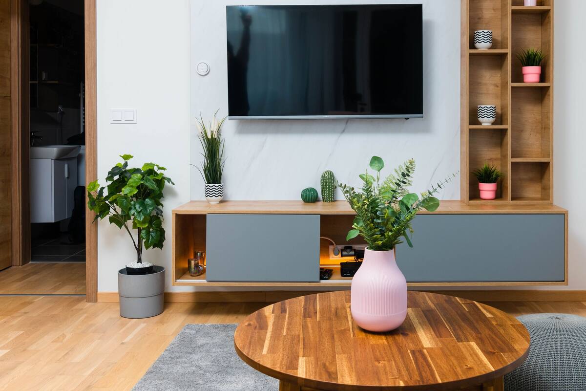 A modern living room with a wooden floor, a round wooden table, a pink vase, a TV mounted on the wall, and a grey cabinet with plants.