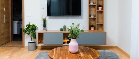 A modern living room with a wooden floor, a round wooden table, a pink vase, a TV mounted on the wall, and a grey cabinet with plants.