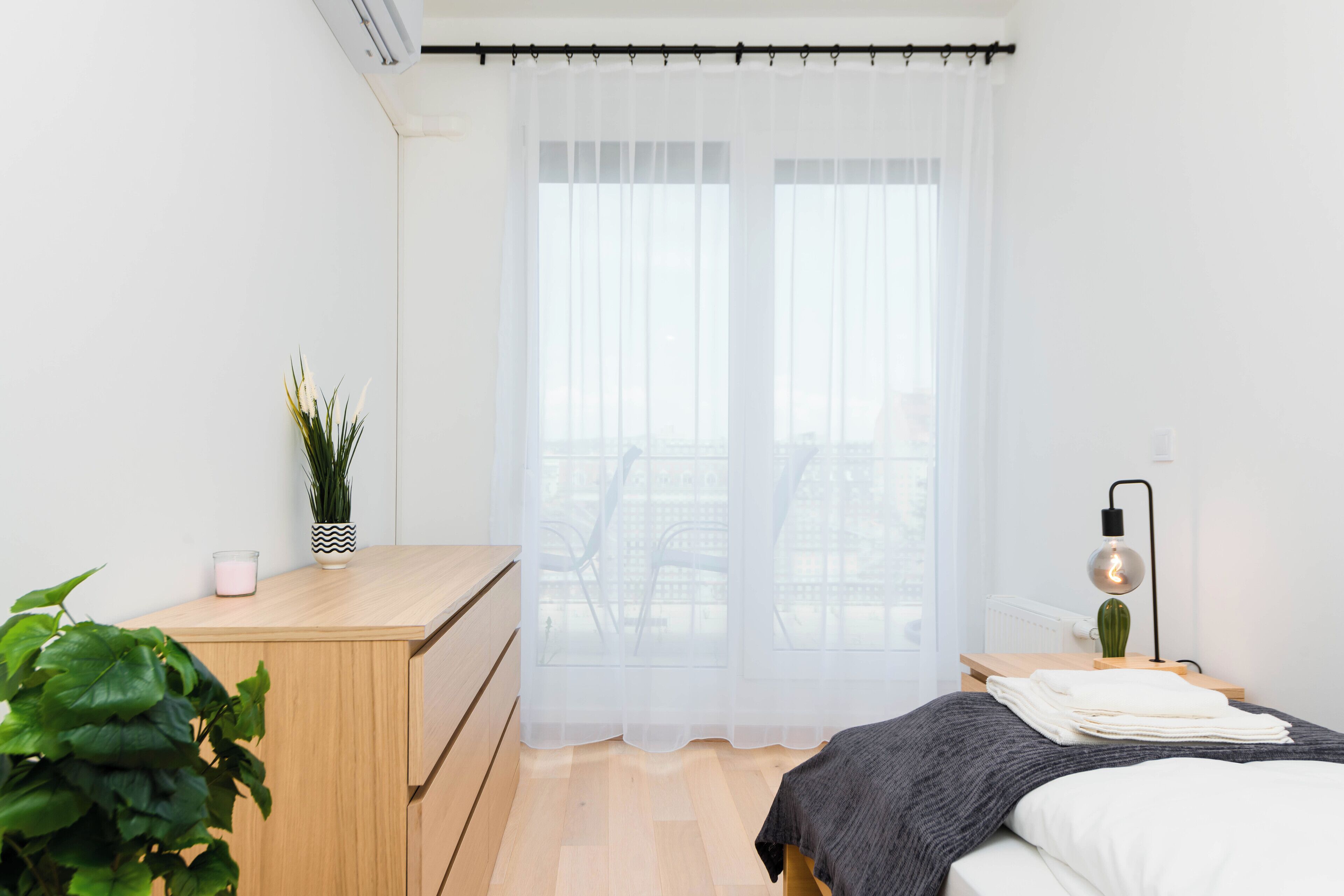 Une chambre moderne avec un bureau en bois, un lit recouvert d’une couverture blanche et grise, une petite plante et une lampe.