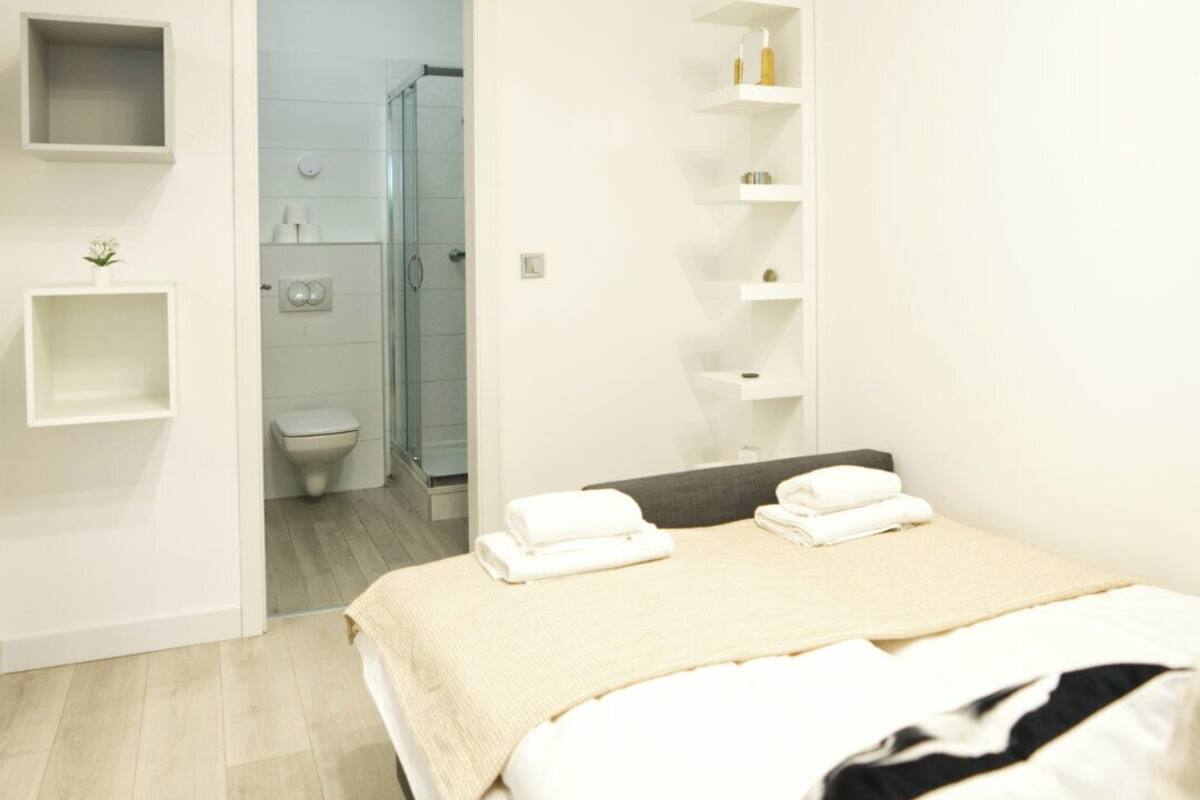A modern bedroom with a bed, a bedside table, a built-in shelf unit, and a view into the bathroom.