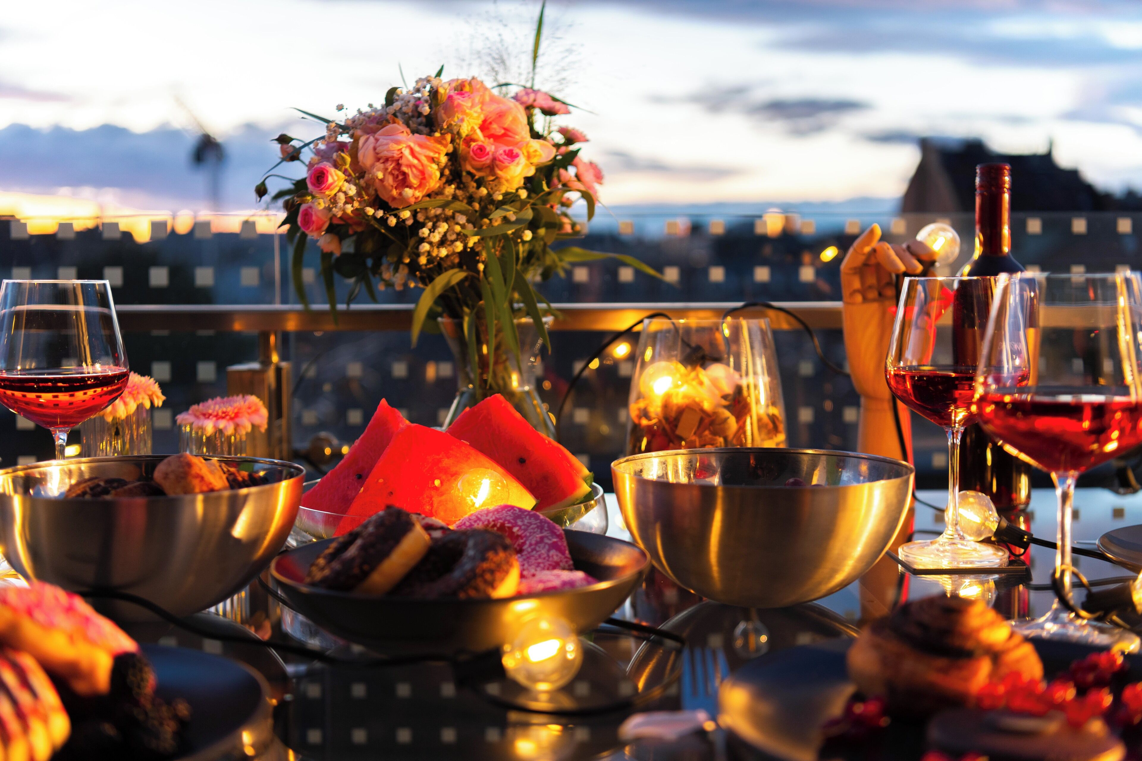 A table set with various dishes, glasses of red wine, and a bouquet of flowers against a cityscape backdrop.