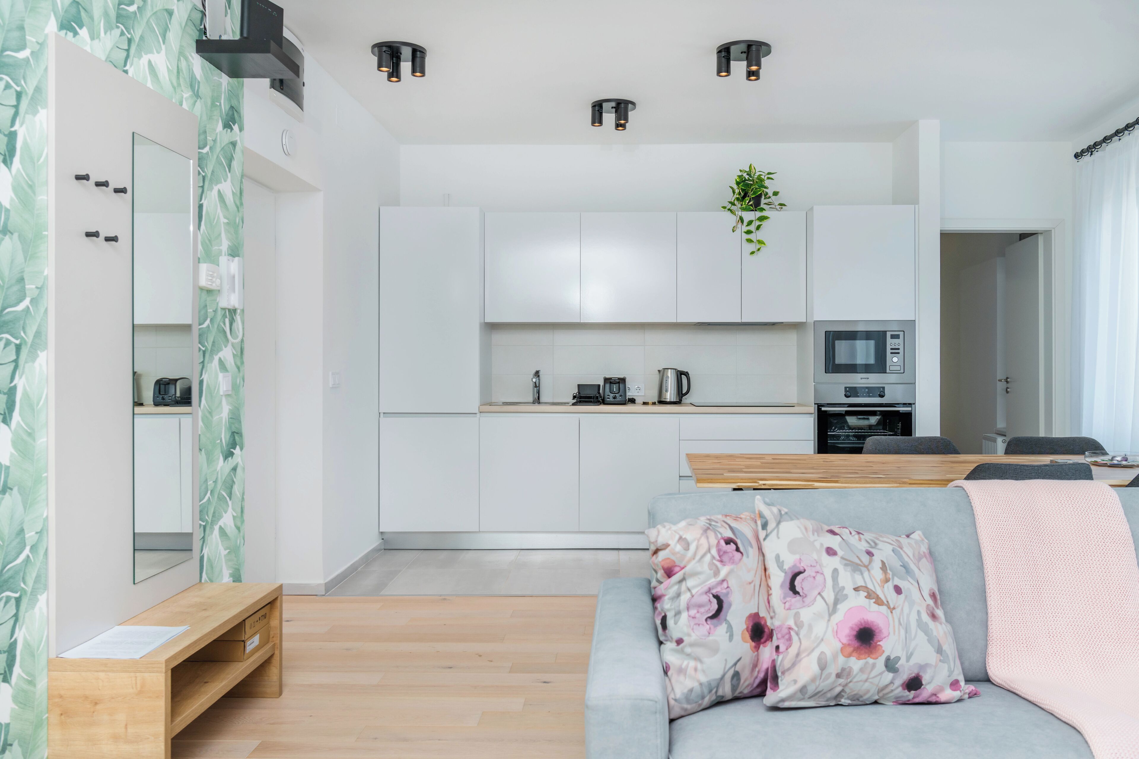 A modern living space with a grey sofa, wooden side table, and a kitchen area with white cabinets and appliances.