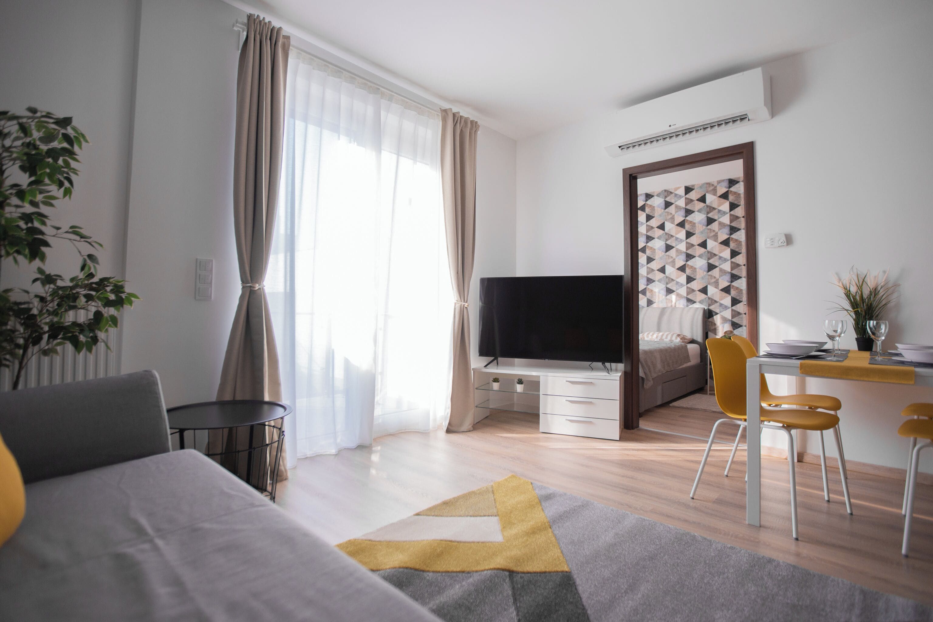 A modern hotel room with a bed, a desk with a computer, a TV, and a dining area with chairs and a table.