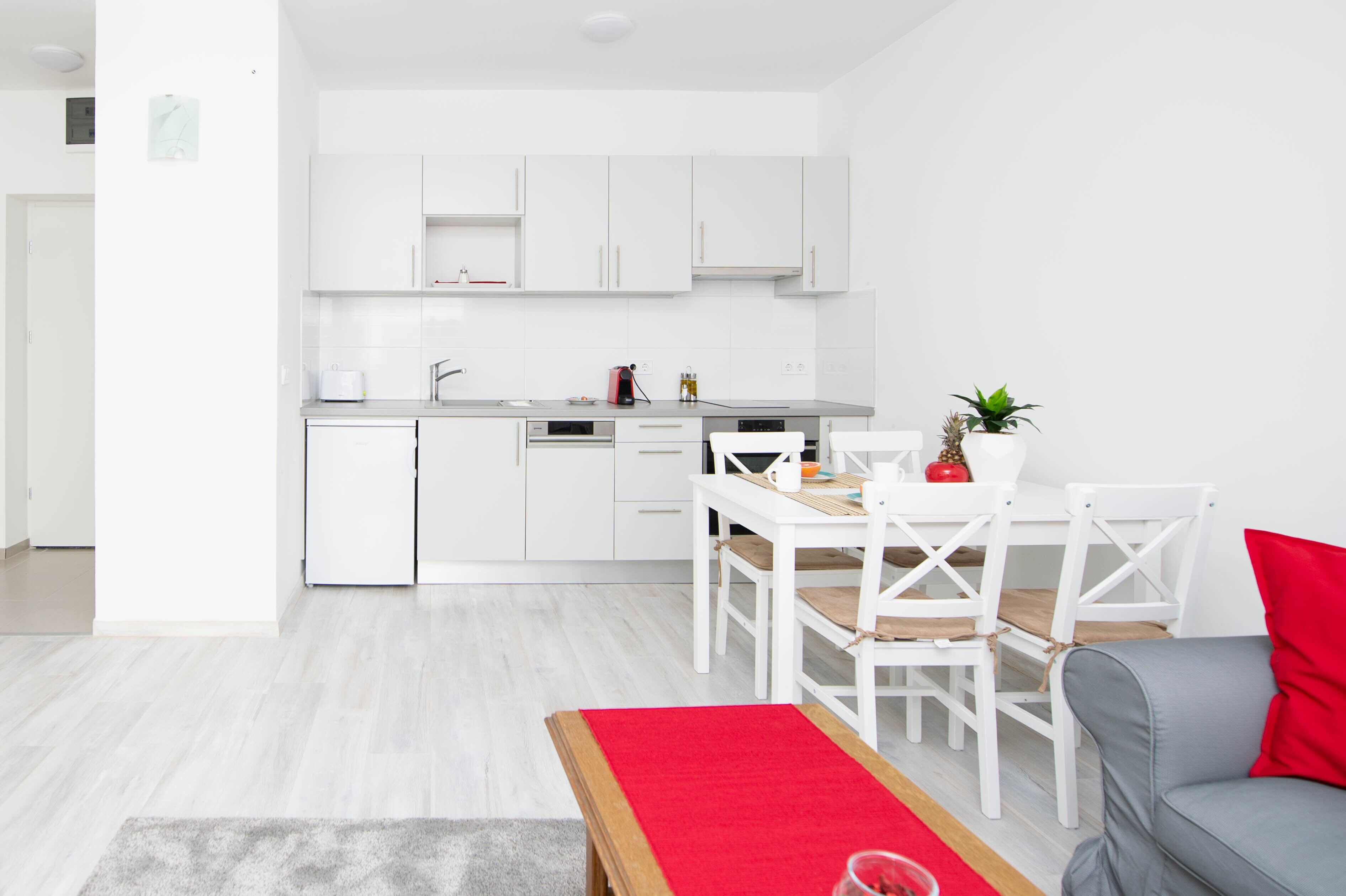 A modern kitchen with white cabinets, a dining table with chairs, and a grey sofa with a red pillow.