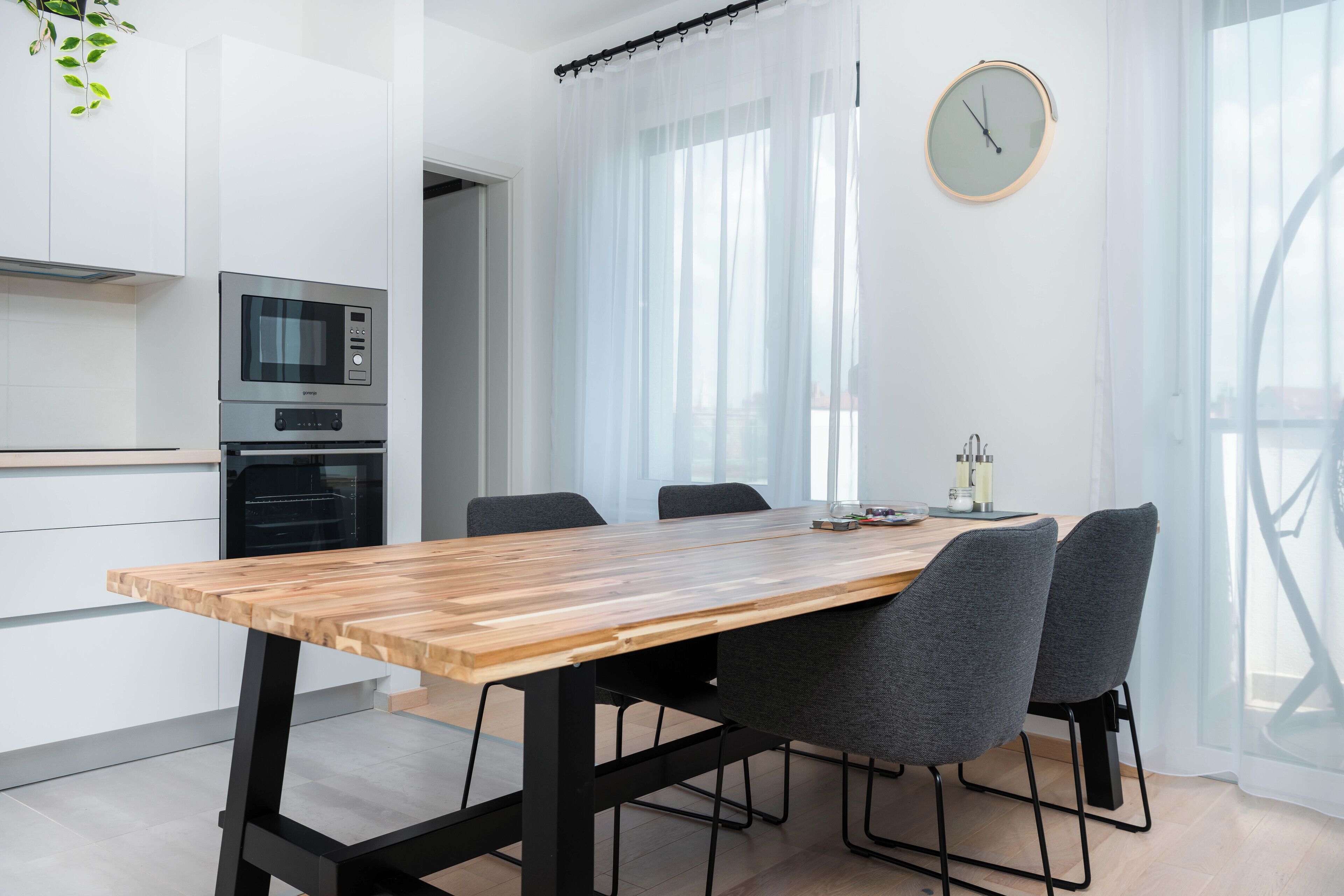 A modern kitchen with a wooden dining table, two grey chairs, and a clock on the wall.