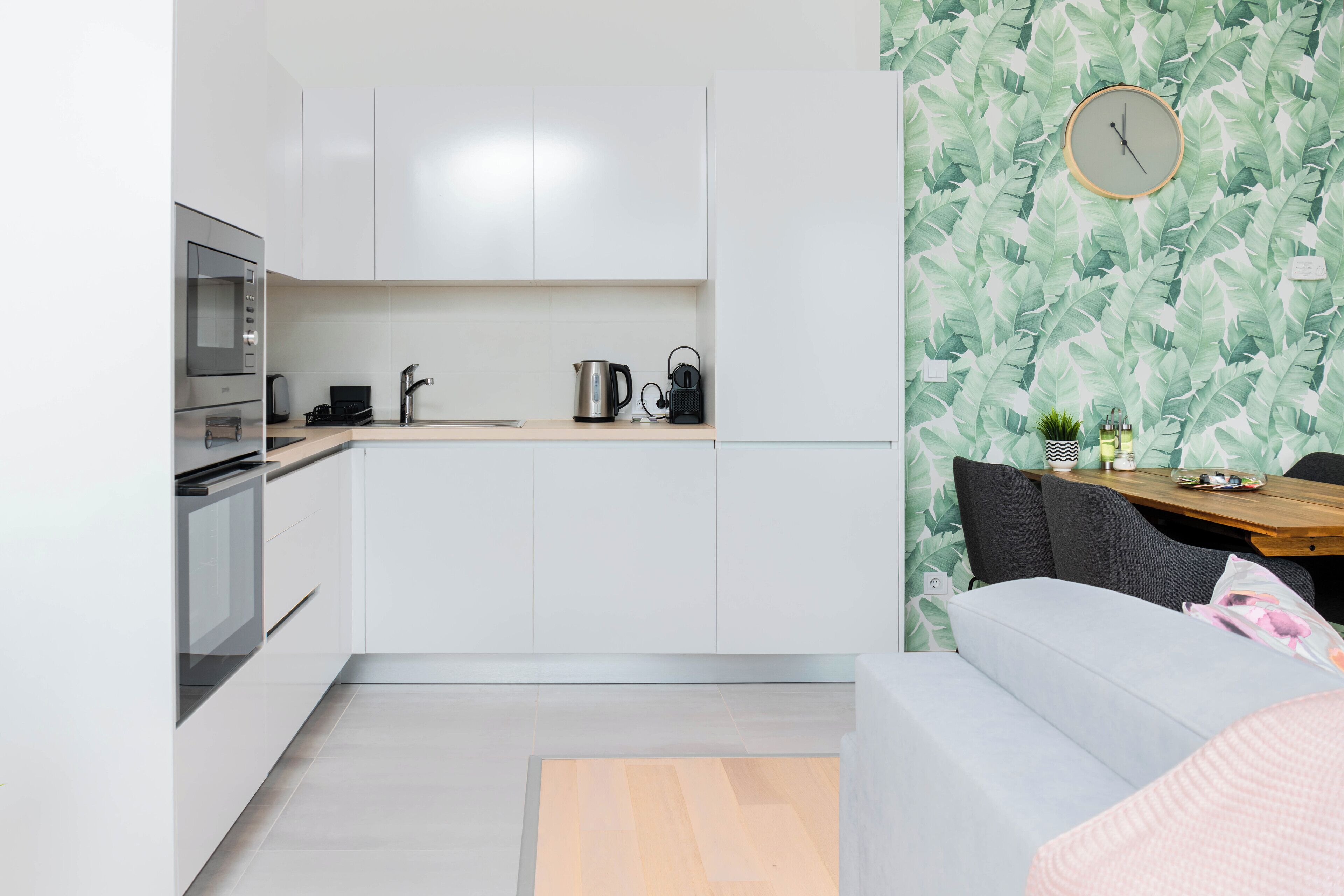 Een moderne keuken met witte kastjes, een magnetron en een waterkoker. Er staat een eettafel met stoelen en een klok hangt aan de muur.