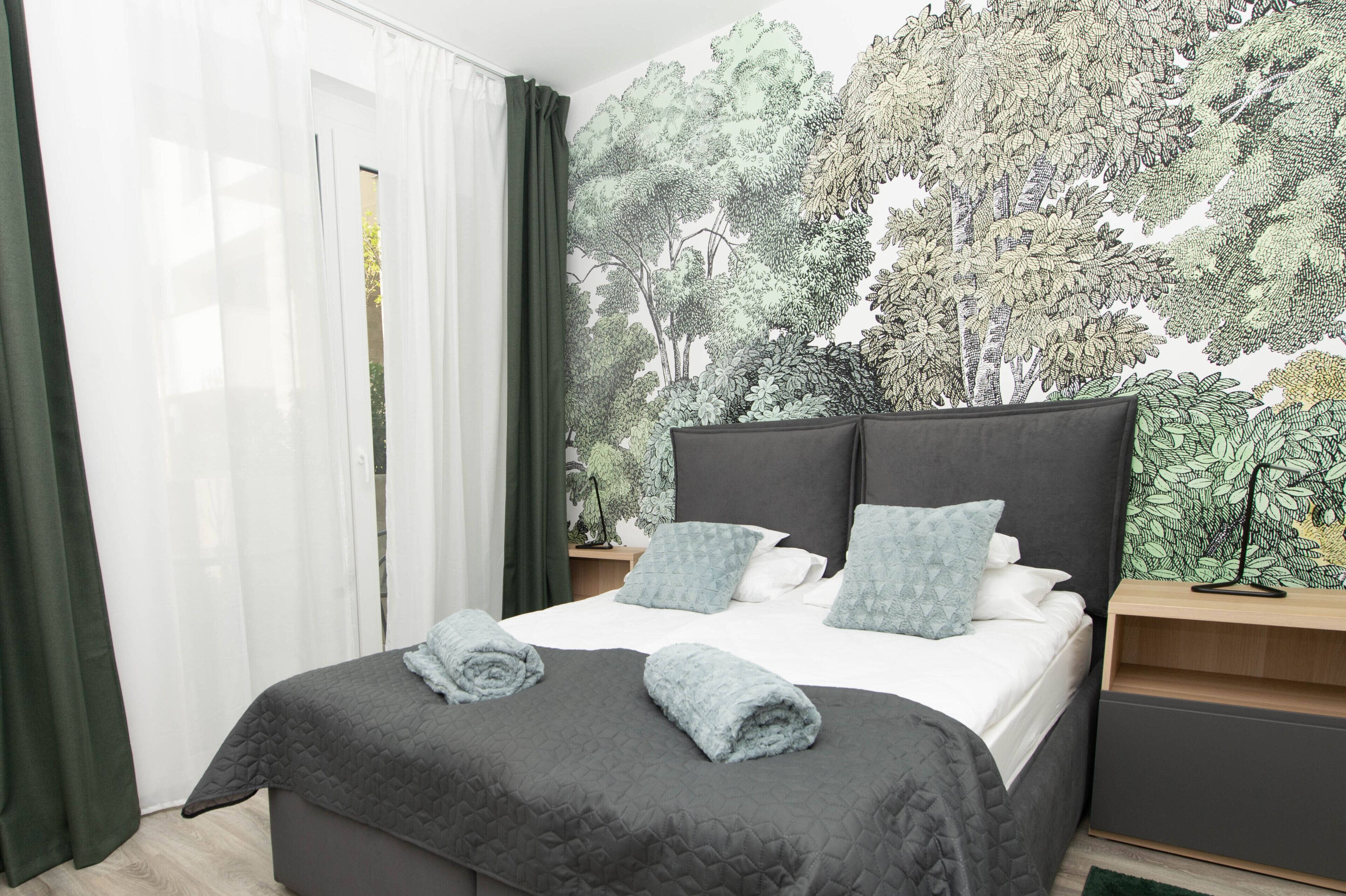 A bedroom with a bed, green curtains, a wall mural of trees, and a nightstand.