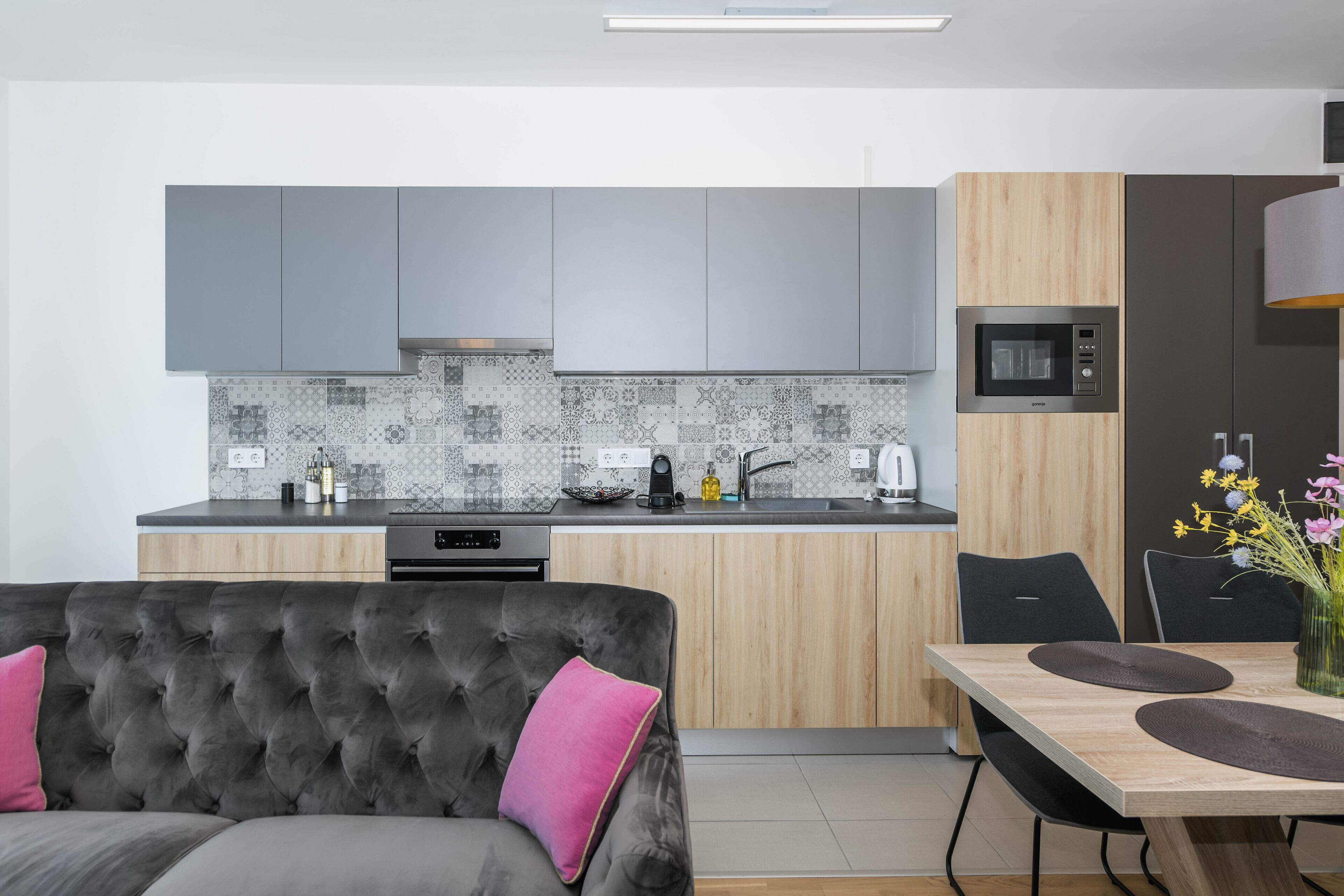 Una cocina moderna con armarios grises, salpicadero de azulejos y comedor con mesa y sillas de madera.