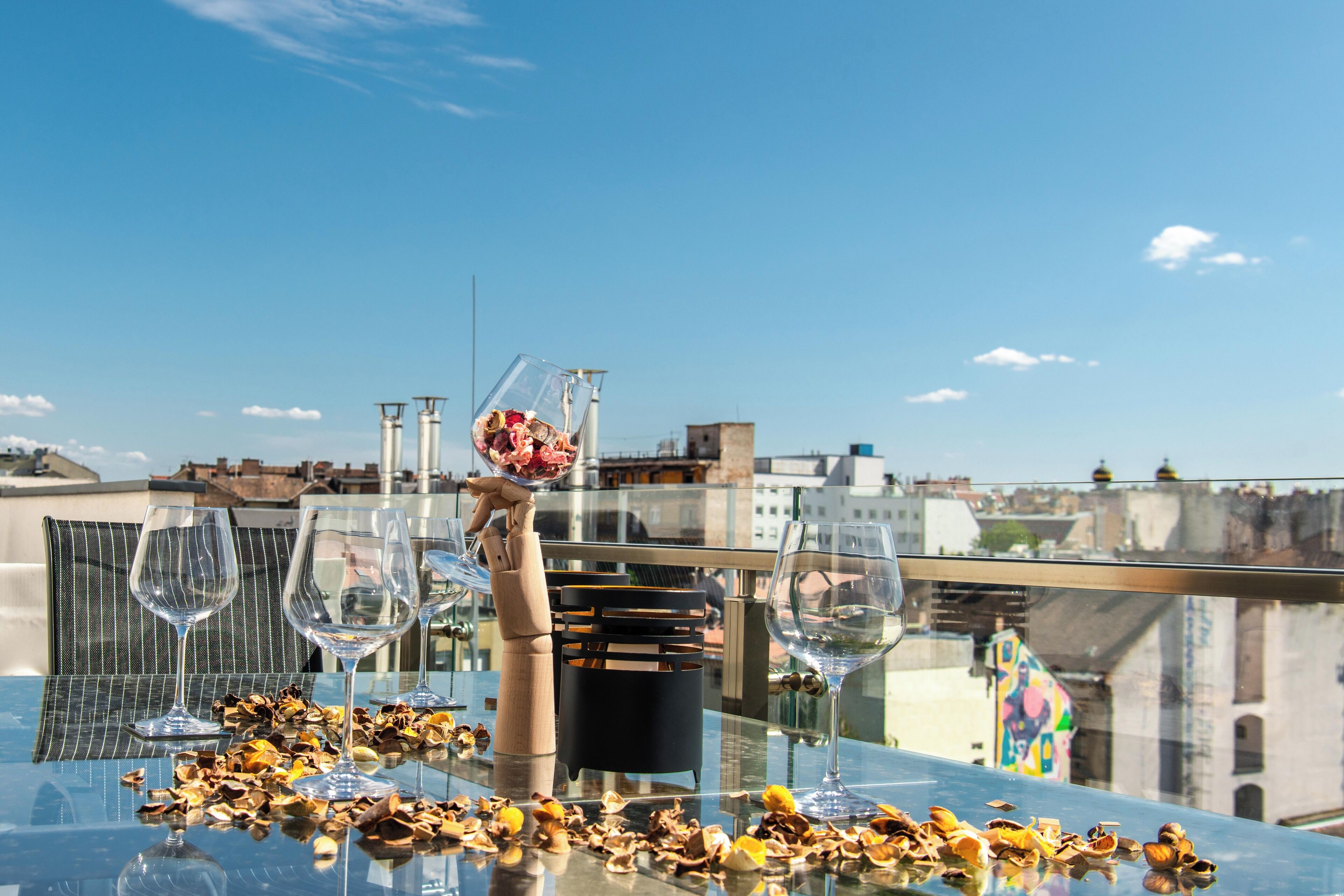 A glass table with wine glasses and a bowl of flowers on a rooftop terrace with a city view.