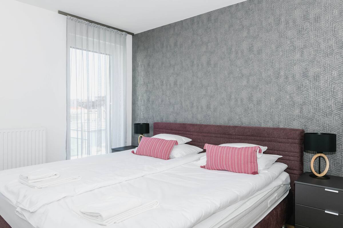 A neatly made bed with white linens and pink striped pillows, a dark headboard, and a nightstand with a lamp.