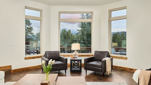 A room with three large windows, a wooden floor, two leather chairs, a small table with a lamp, and a vase with flowers.