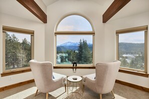 A room with a large arched window, two armchairs, a small round table, and a view of a forest and mountains.